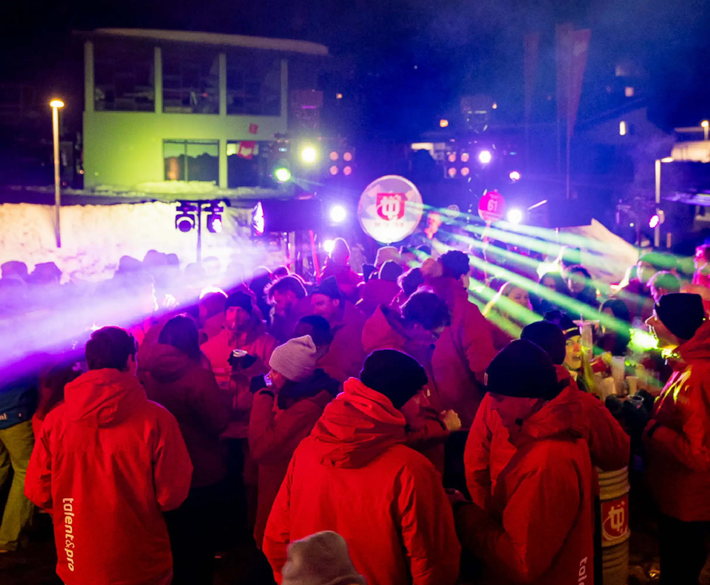 Crowd of young people gathered outdoors in front of a stage with performers and colorful DBSF banners and lighting.