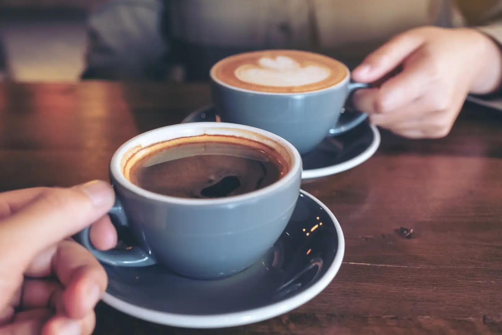 Two hands holding gray cups of coffee over a wooden table; one cup holds black coffee, the other a latte with heart art. Cozy, inviting atmosphere.