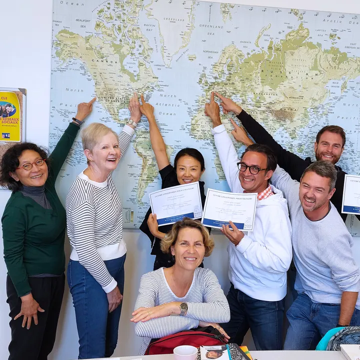 A group of six students and their instructor, Emmanuelle, pointing at their home cities on a large map on the wall.