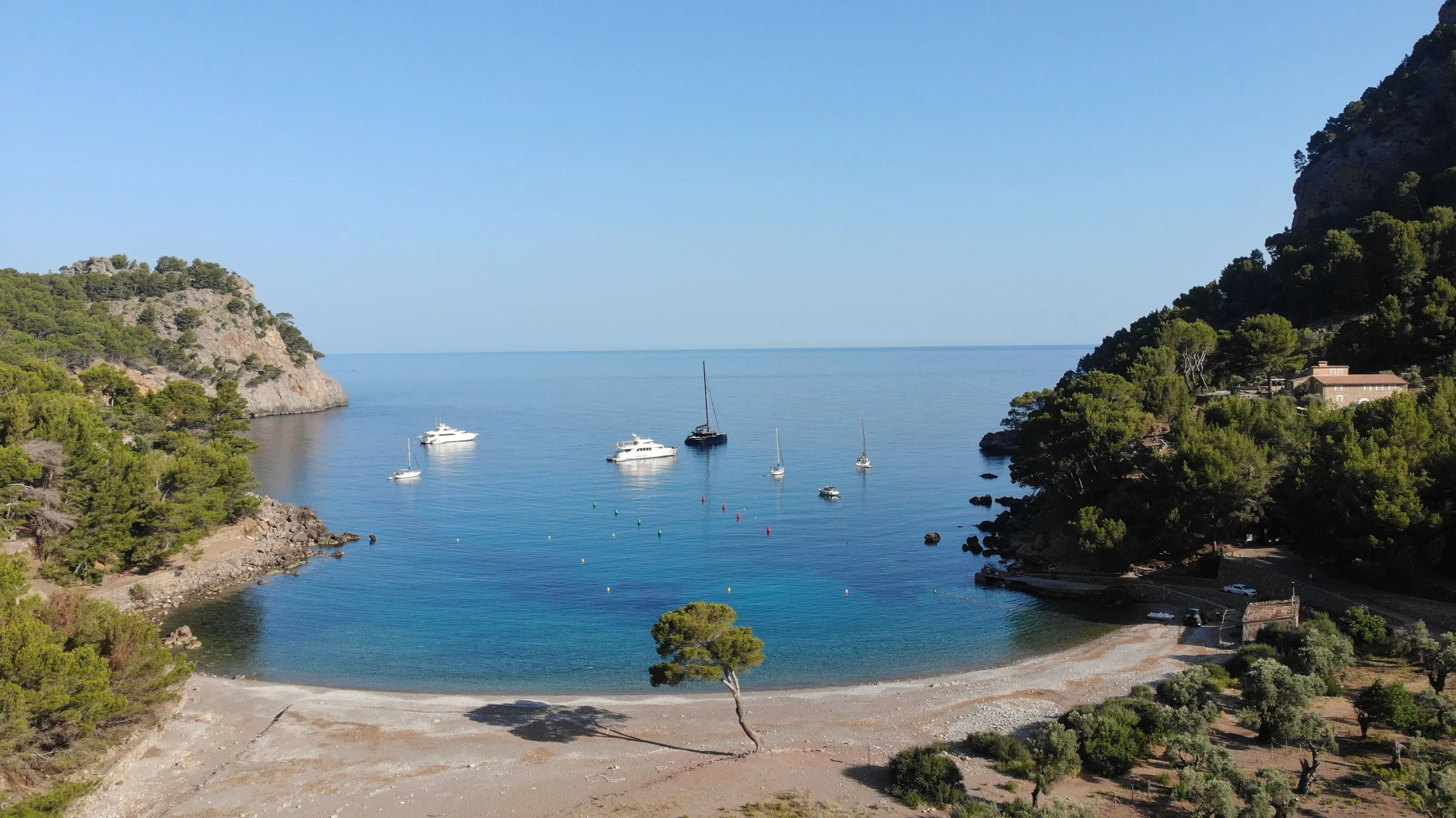 Calm bay at Cala Tuent with anchored sailboats and a quiet beach curving between forested headlands.
