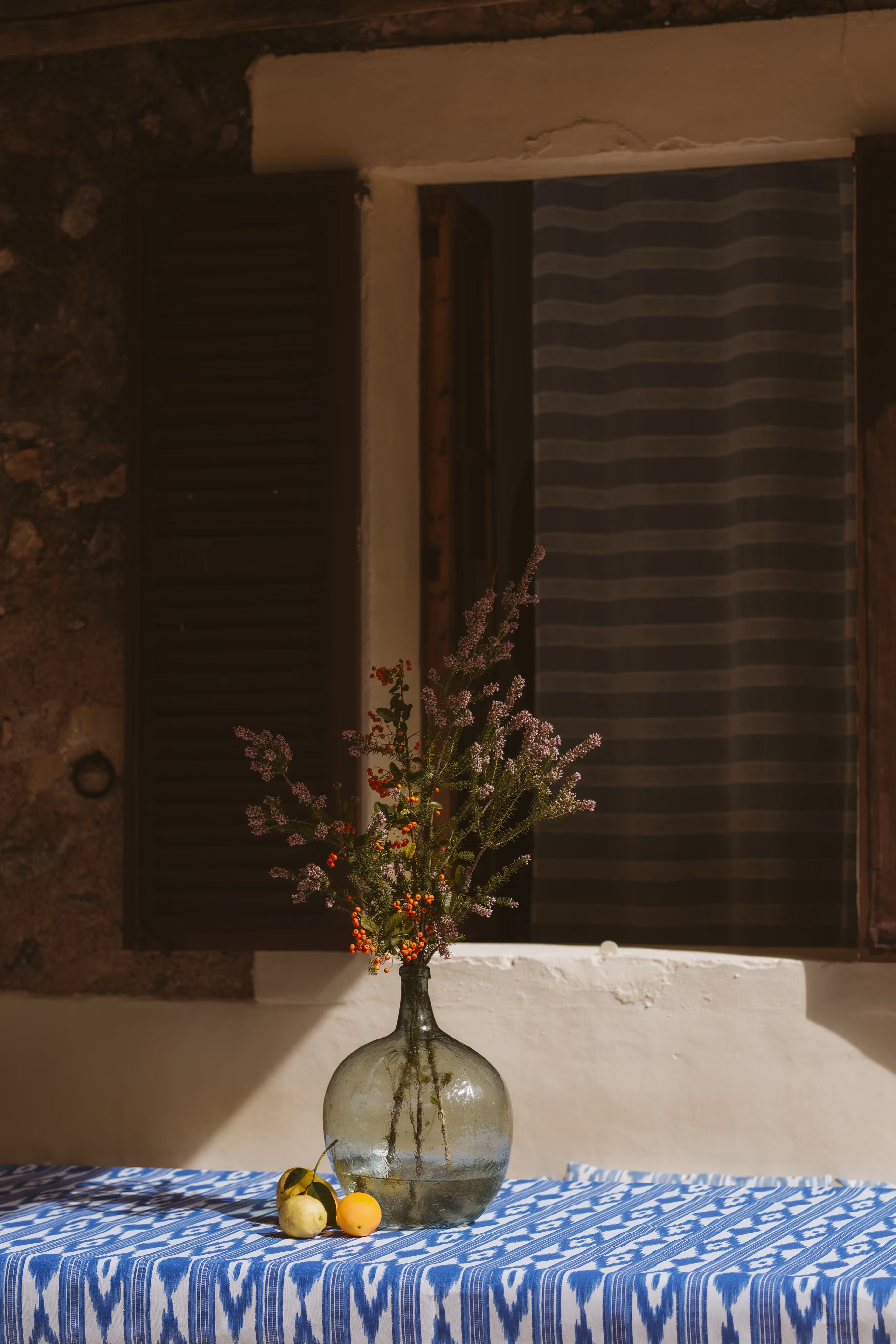 Glass vase with wildflowers on a rustic table with lemons, set against a sunlit wall and wooden shutters at a Mediterranean farmhouse.