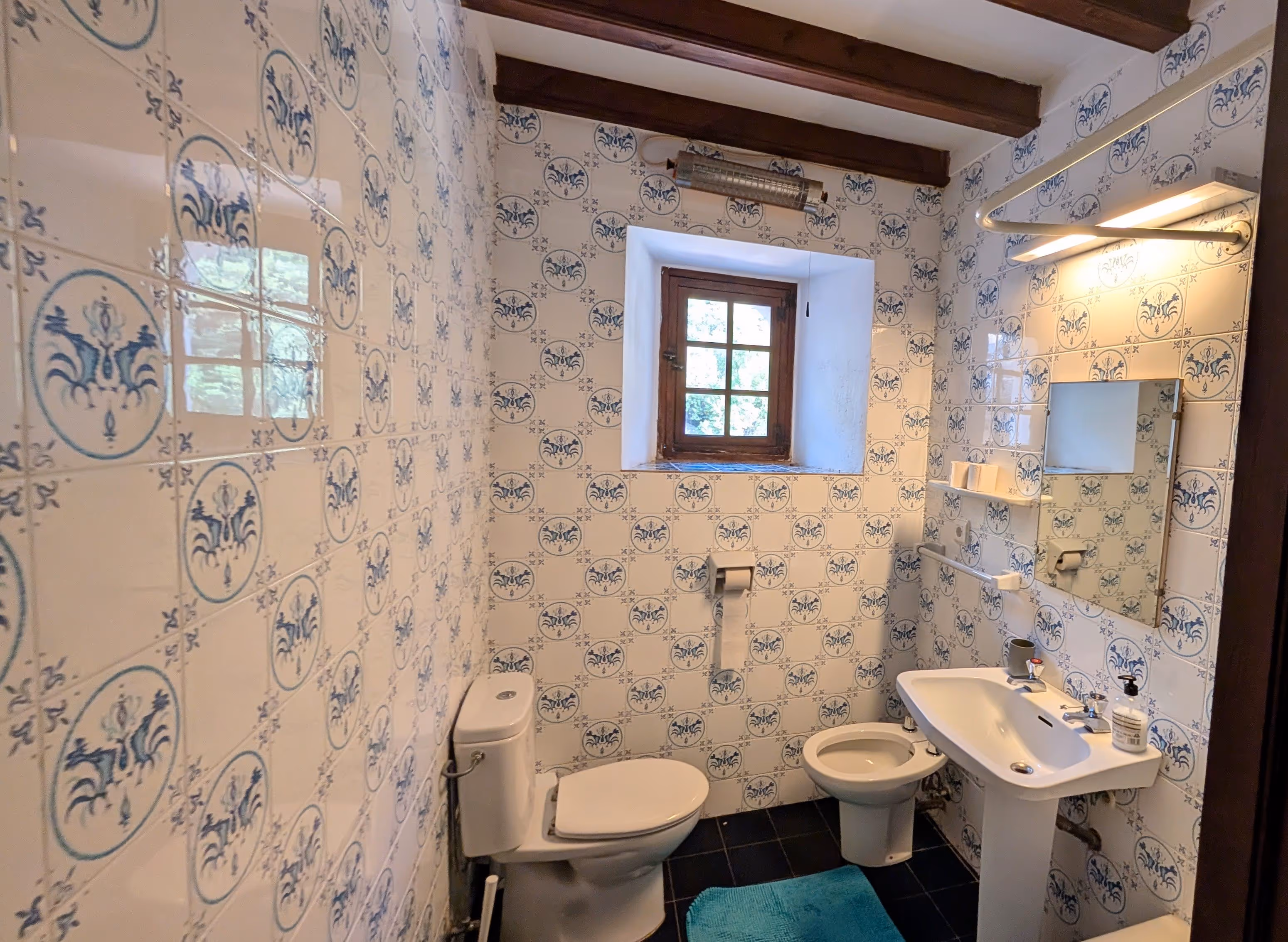 Blue-and-white tiled bathroom with a small window, toilet, bidet, and sink in a rustic setting.