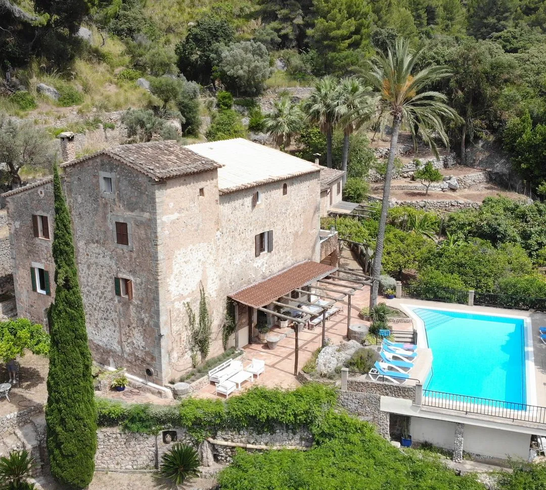 Aerial view of Finca Higo’s traditional stone house with a shaded terrace, sun loungers, and a bright blue pool surrounded by lush greenery and palm trees in the Tramuntana mountains.