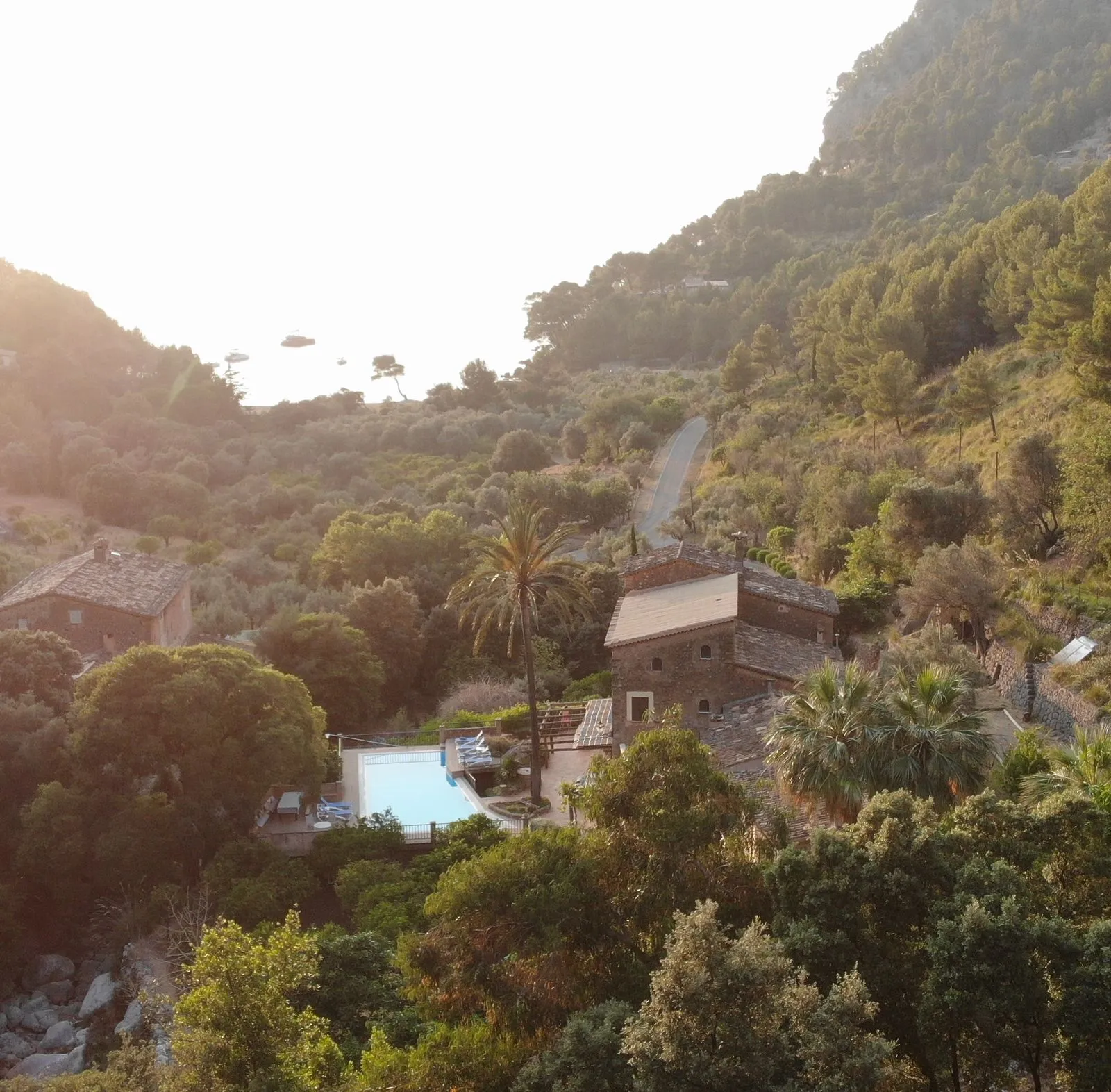 View from Finca Higo overlooking a lush green valley with terraced stone houses, a pool, and a distant view of the beach and sea at sunset.