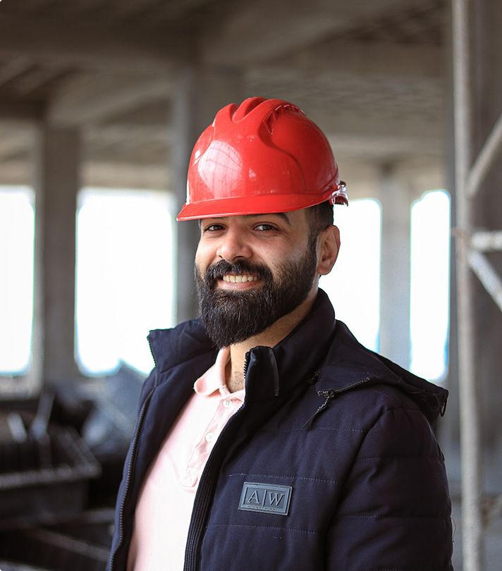 Smiling man with beard wearing a red hard hat and dark jacket at a construction site.
