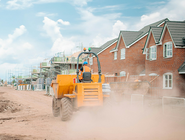 Worker operating yellow construction vehicle on dusty road by houses under construction.