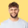 Smiling man with curly hair and beard wearing a light blue t-shirt against a yellow background.