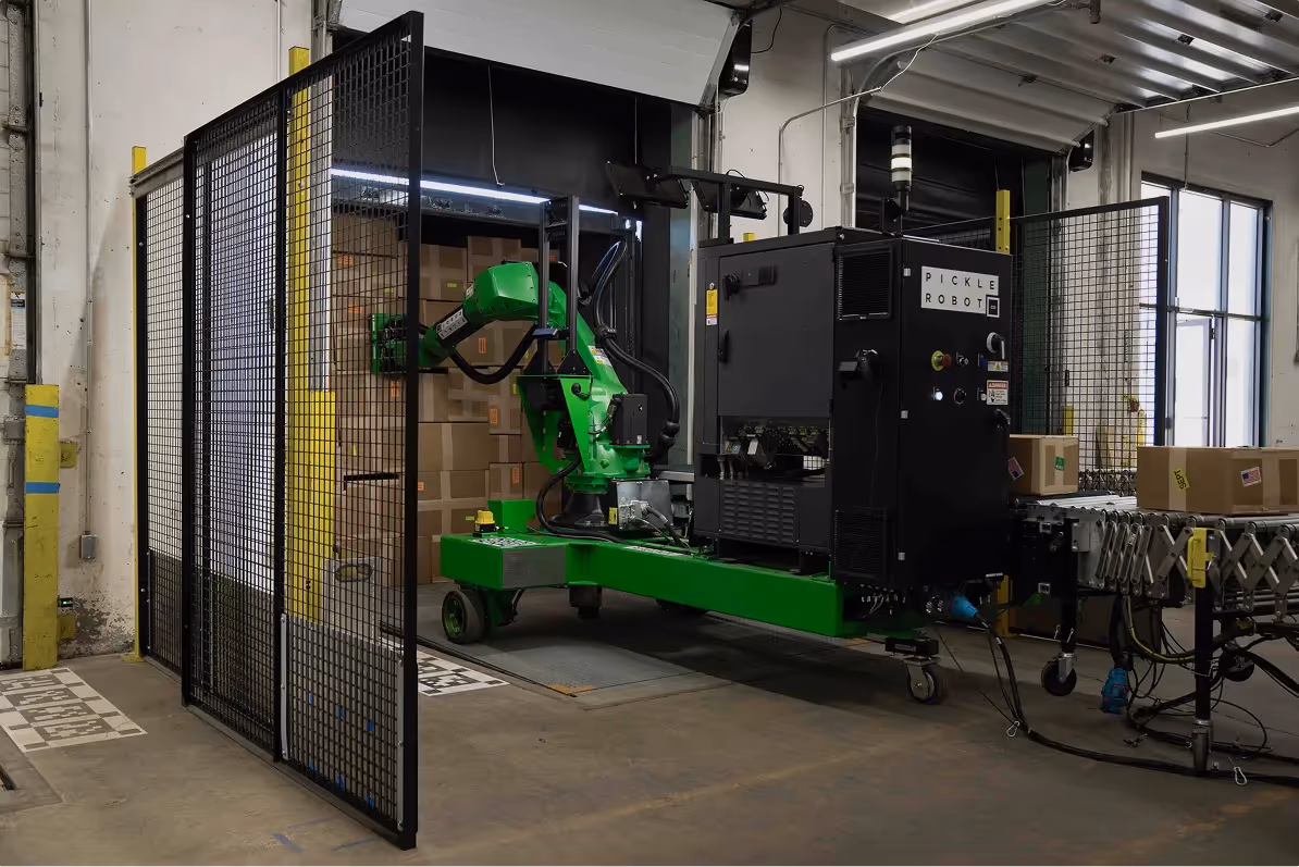 Green robotic arm labeled Pickle Robot working inside a gated industrial packaging area with brown boxes on conveyor belts.