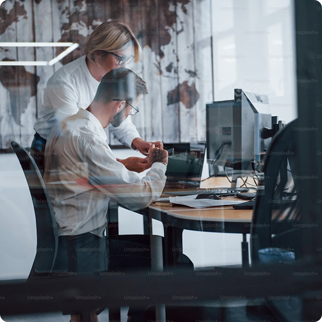Two colleagues working together at an office desk with computer monitors; a woman is standing and showing something to a seated man.