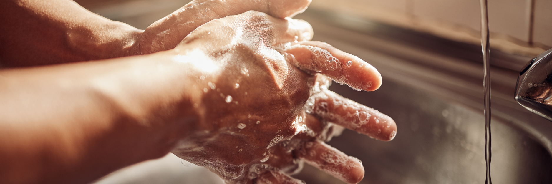 Close-up of hands covered with soap suds under running water in a sink, demonstrating hand washing.