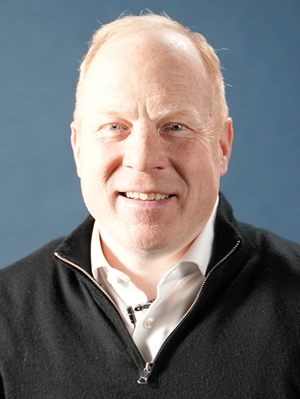 Smiling Mike Smith man with light skin, short light hair, wearing a black zip-up sweater over a white collared shirt against a blue background.