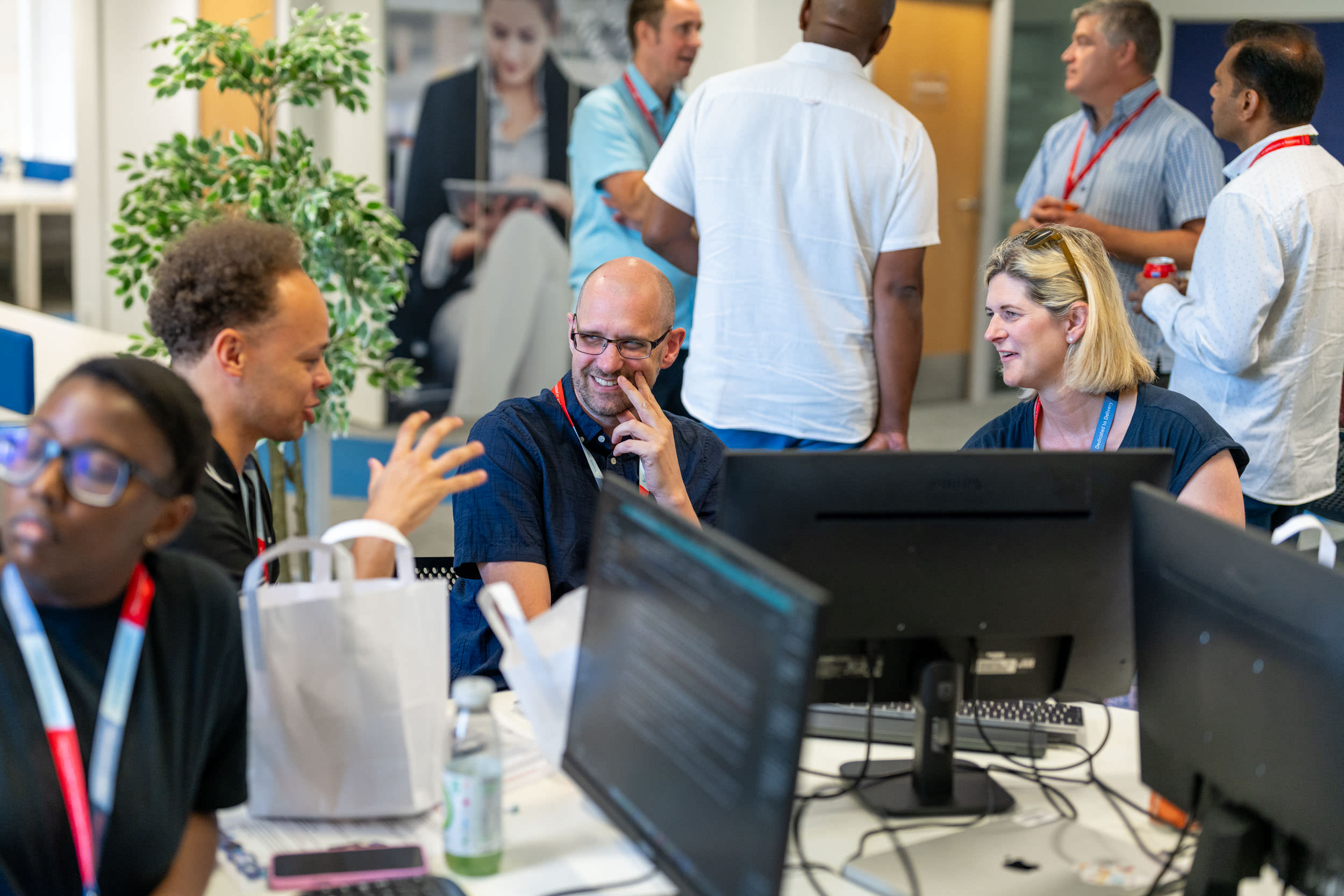 A group of office workers engage in conversation and collaboration around a desk with computer monitors in a modern office space, with some people talking standing and others seated.