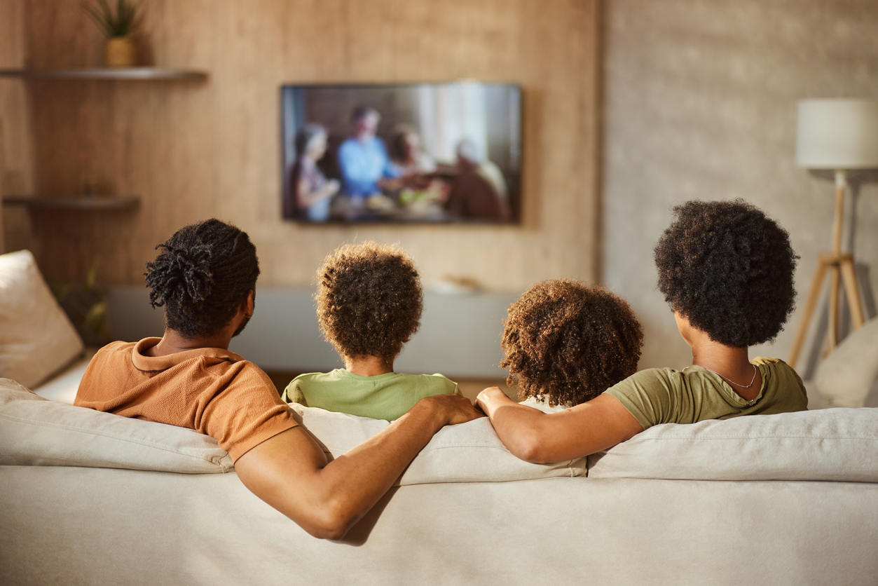 Family of four with curly hair sitting on a beige couch watching TV in a cozy living room.