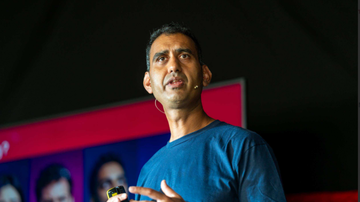 Shuja Khan wearing a blue shirt and a headset microphone speaking or presenting, holding a device in his right hand, with a blurred screen showing people in the background.