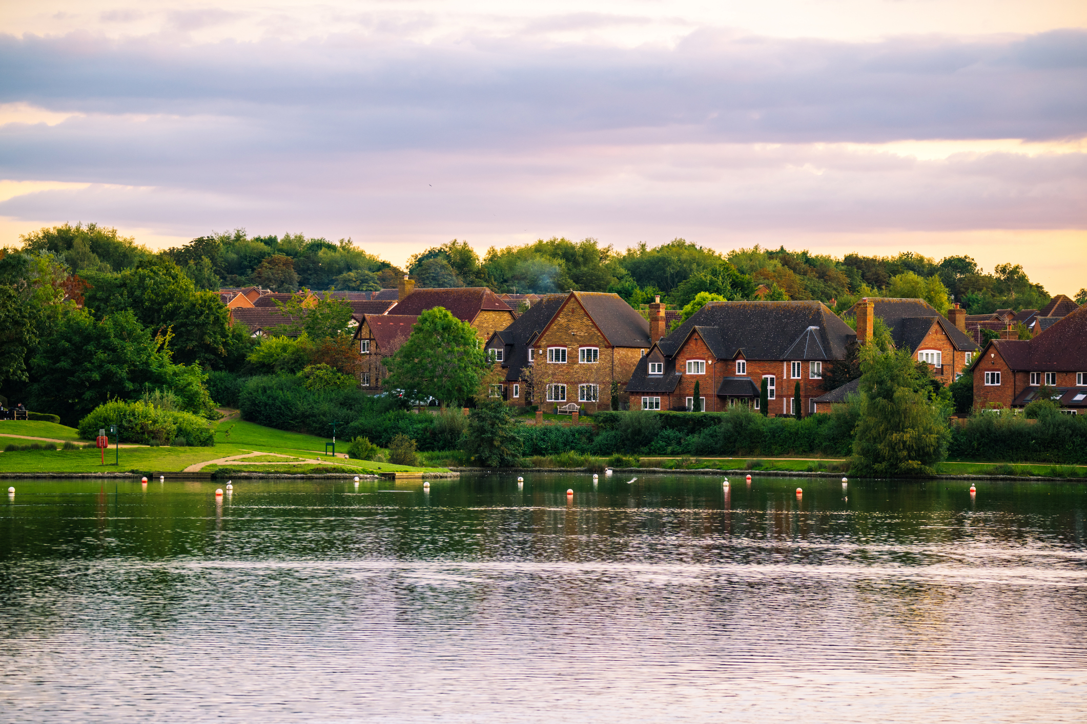 A peaceful lakeside scene with calm water in the foreground, white and red buoys floating, green grassy banks, and a row of brick houses with pitched roofs backed by trees under a cloudy sky at sunset.