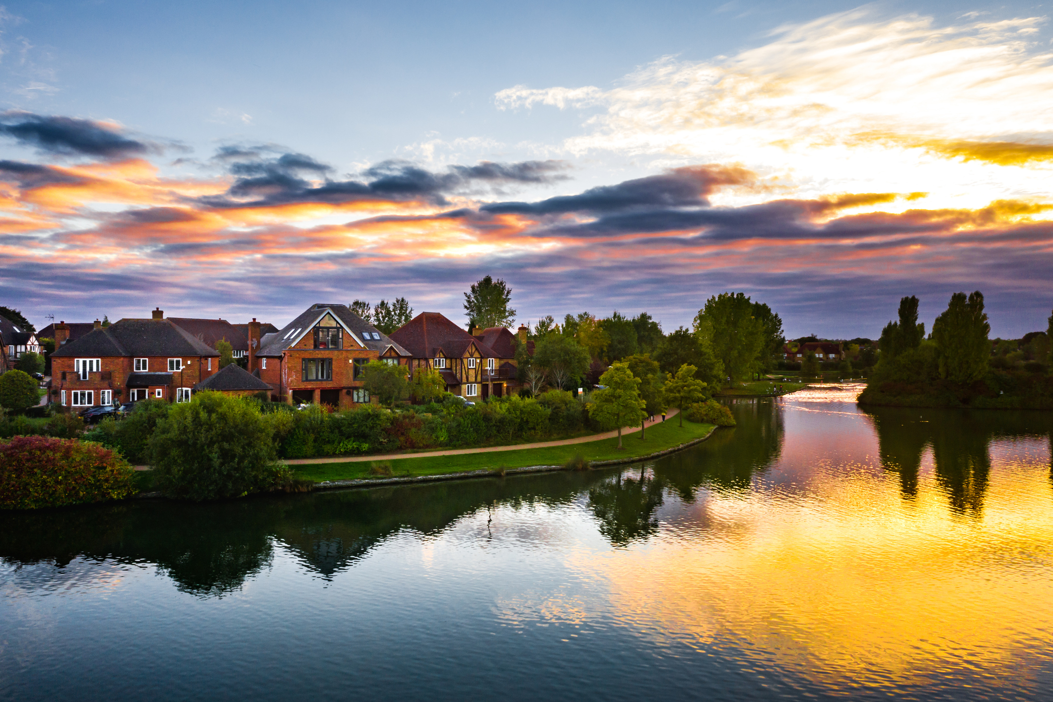 Riverfront houses and trees reflect on the calm water under a colorful sunset sky with orange, purple, and blue clouds.