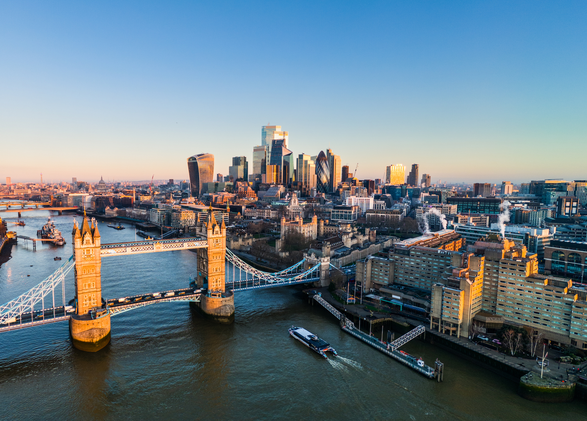 Aerial view of London's Tower Bridge spanning the River Thames with a boat cruising below, and the modern city skyline in the background under a clear blue sky.