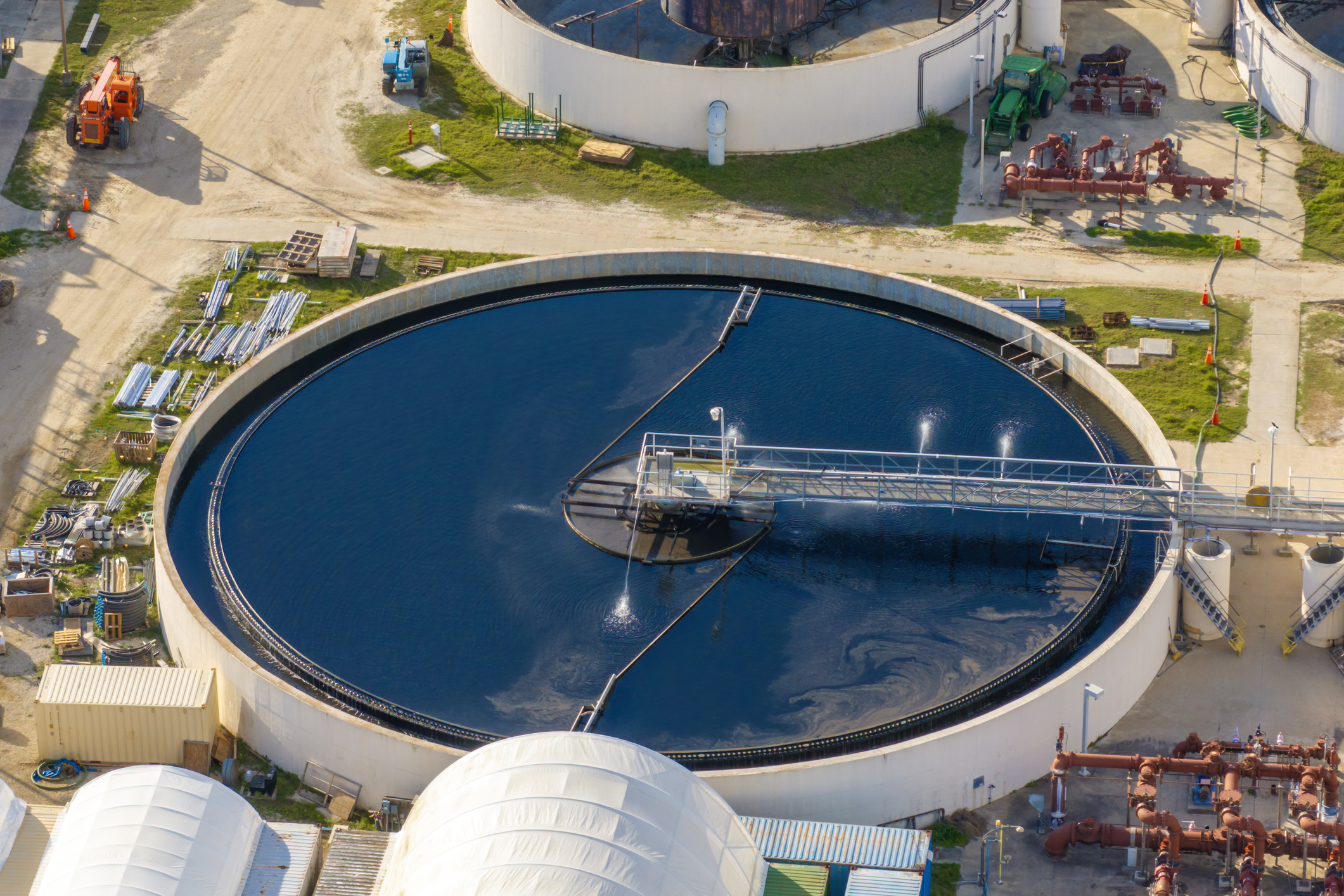 Aerial view of a round water treatment tank with a mechanical arm spraying water inside, surrounded by industrial pipes, equipment, and dirt roads.