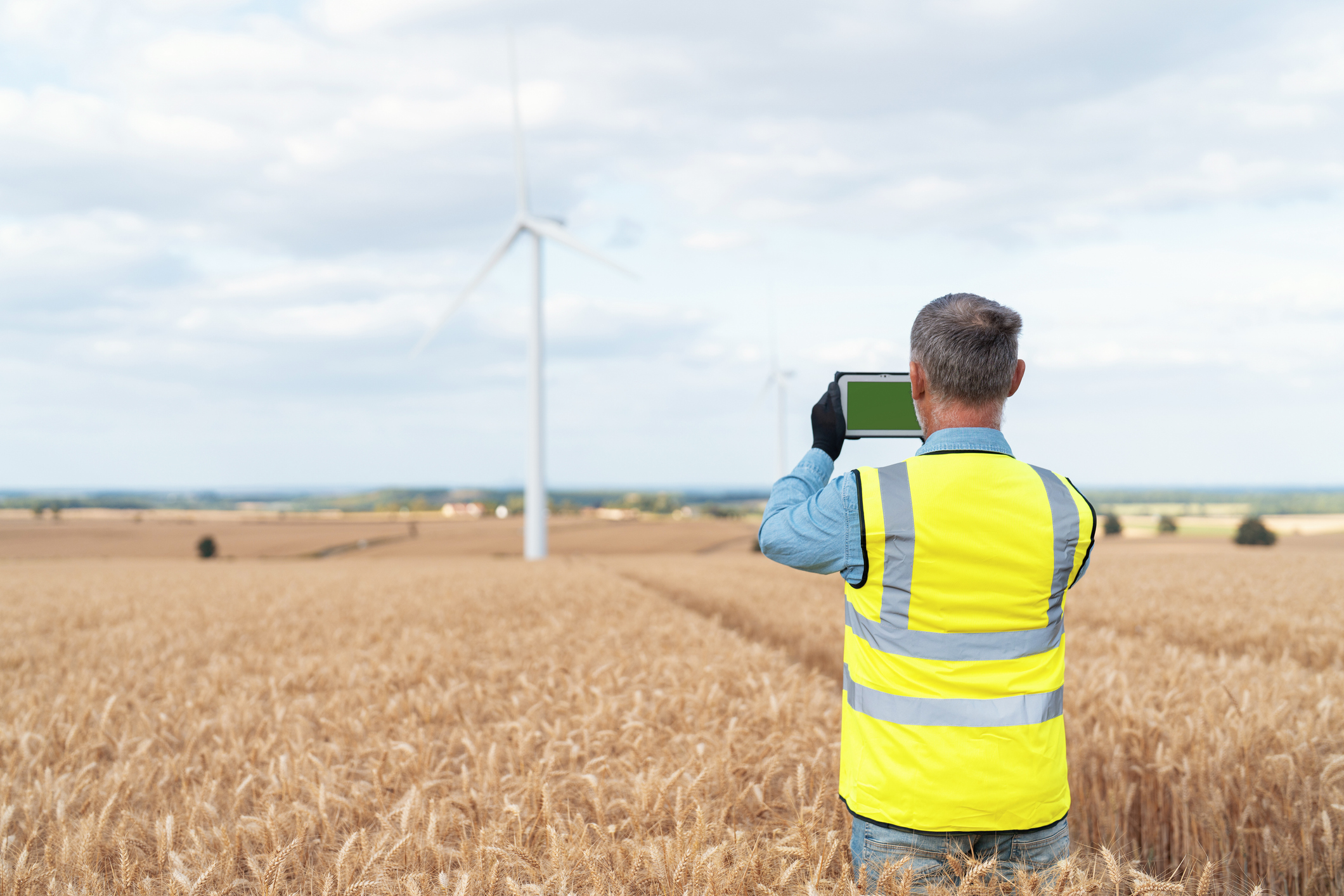 A man wearing a yellow reflective safety vest and black gloves stands in a golden wheat field, holding a tablet with a green screen, facing large wind turbines in the distance under a partly cloudy sky.