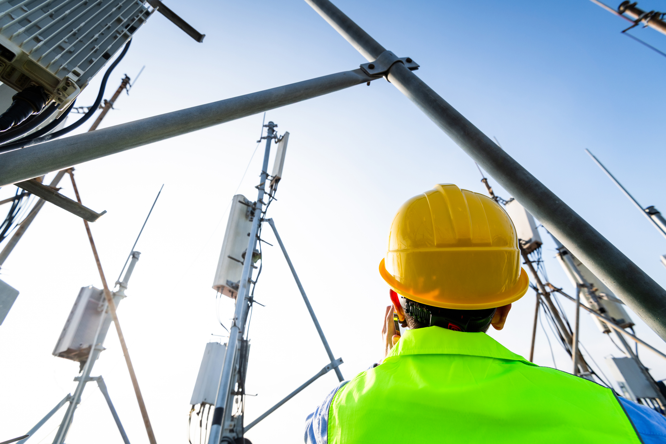 Telecommunication technician wearing a yellow hard hat and neon green safety vest inspecting equipment on a cellular tower against a clear blue sky.