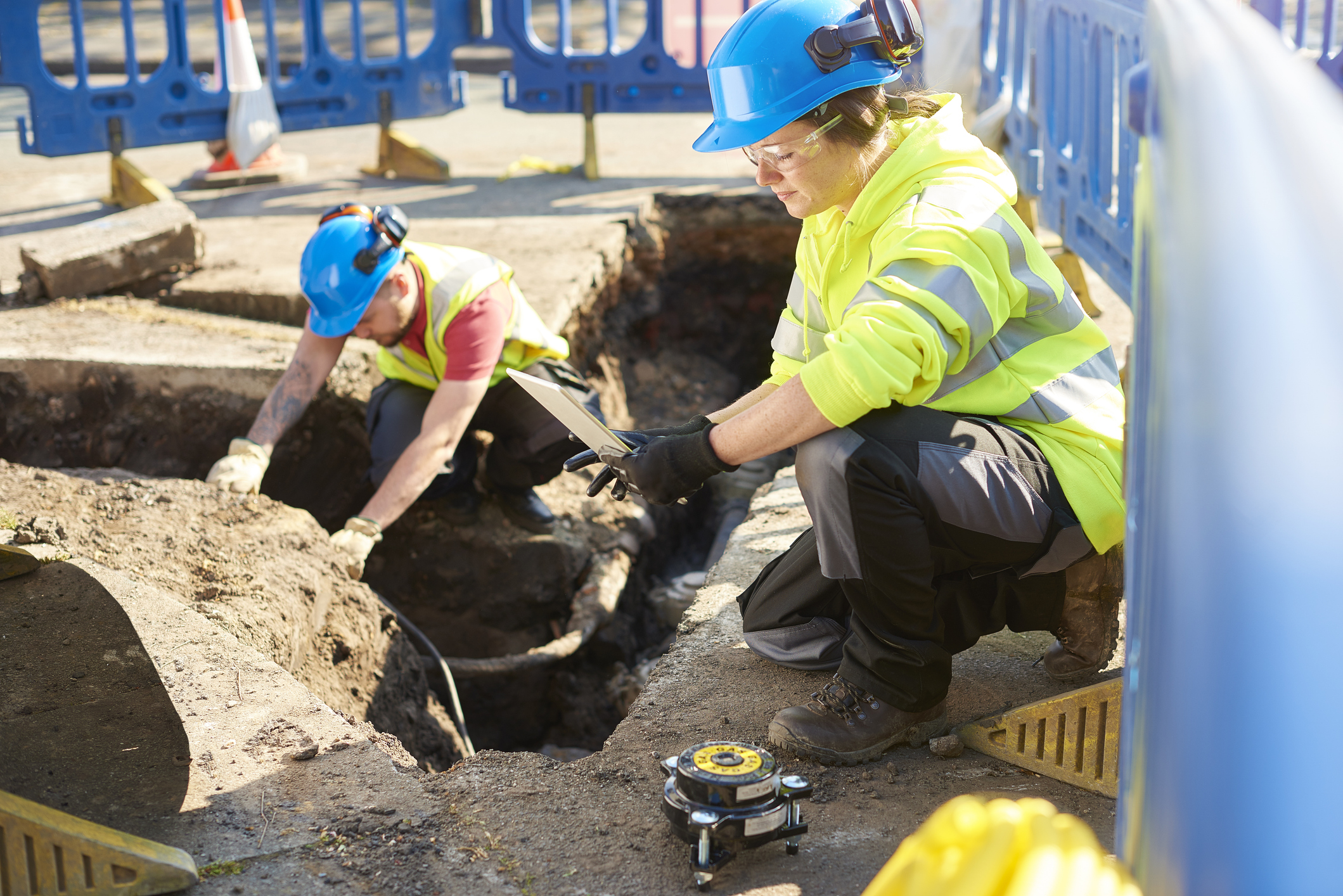 Two construction workers wearing blue helmets and high-visibility clothing working at a road excavation site; one worker is examining the trench while the other is checking a tablet.