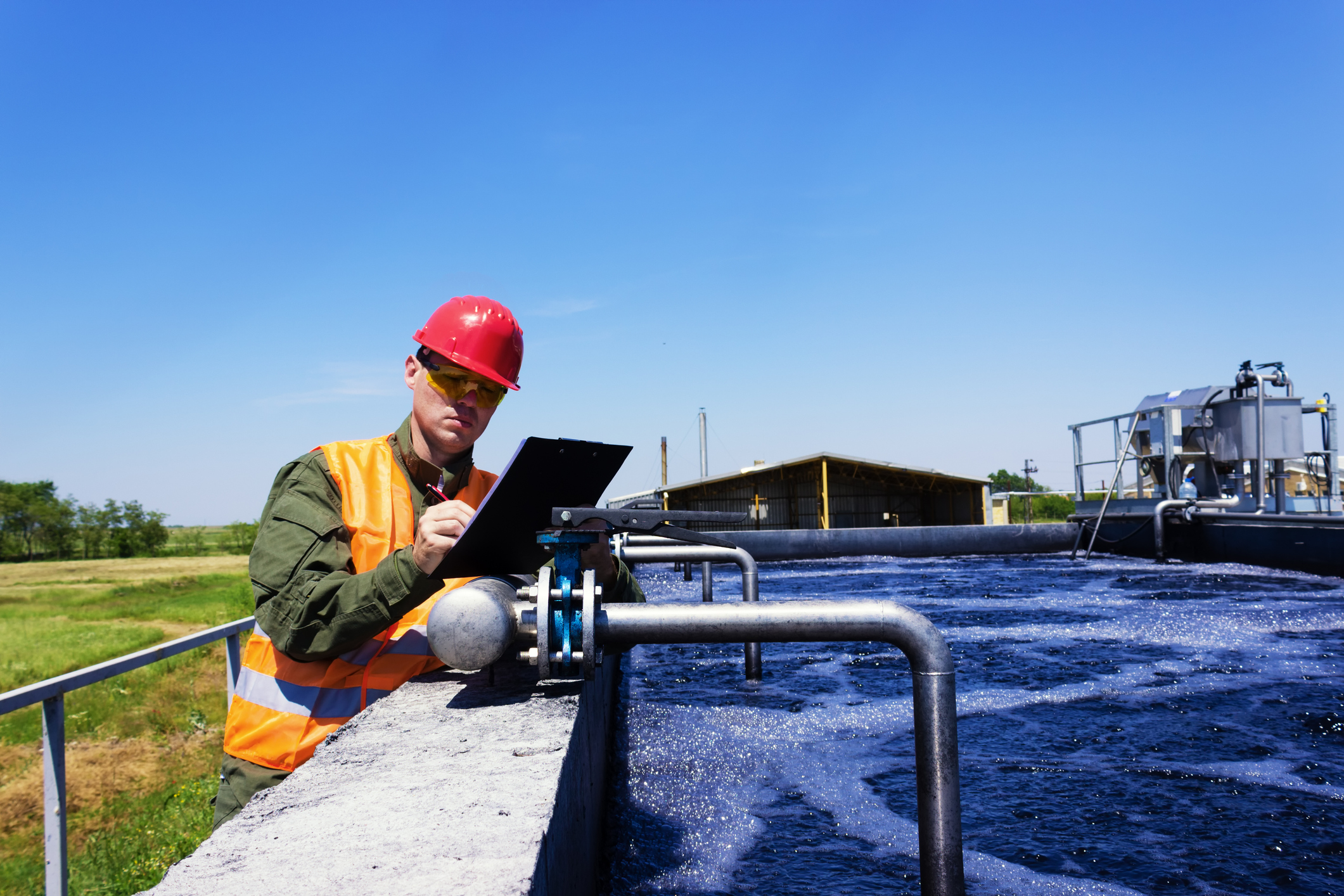 Worker in red hard hat and orange safety vest writing on a clipboard while standing beside an industrial water treatment tank with pipes and aerated water.