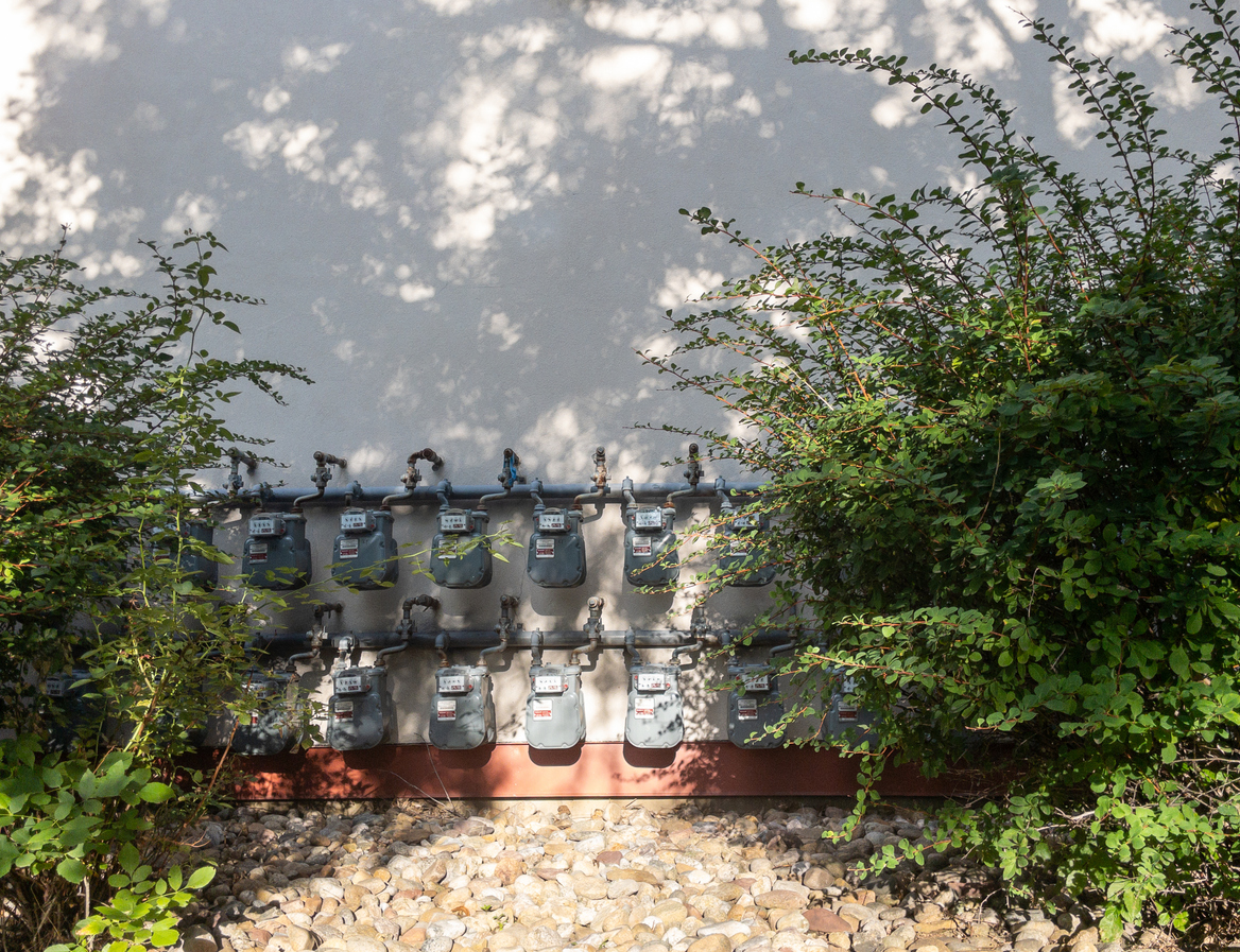 Two rows of gas meters mounted on a light-colored exterior wall between dense green bushes with pebble ground below, dappled by sunlight and shadows.