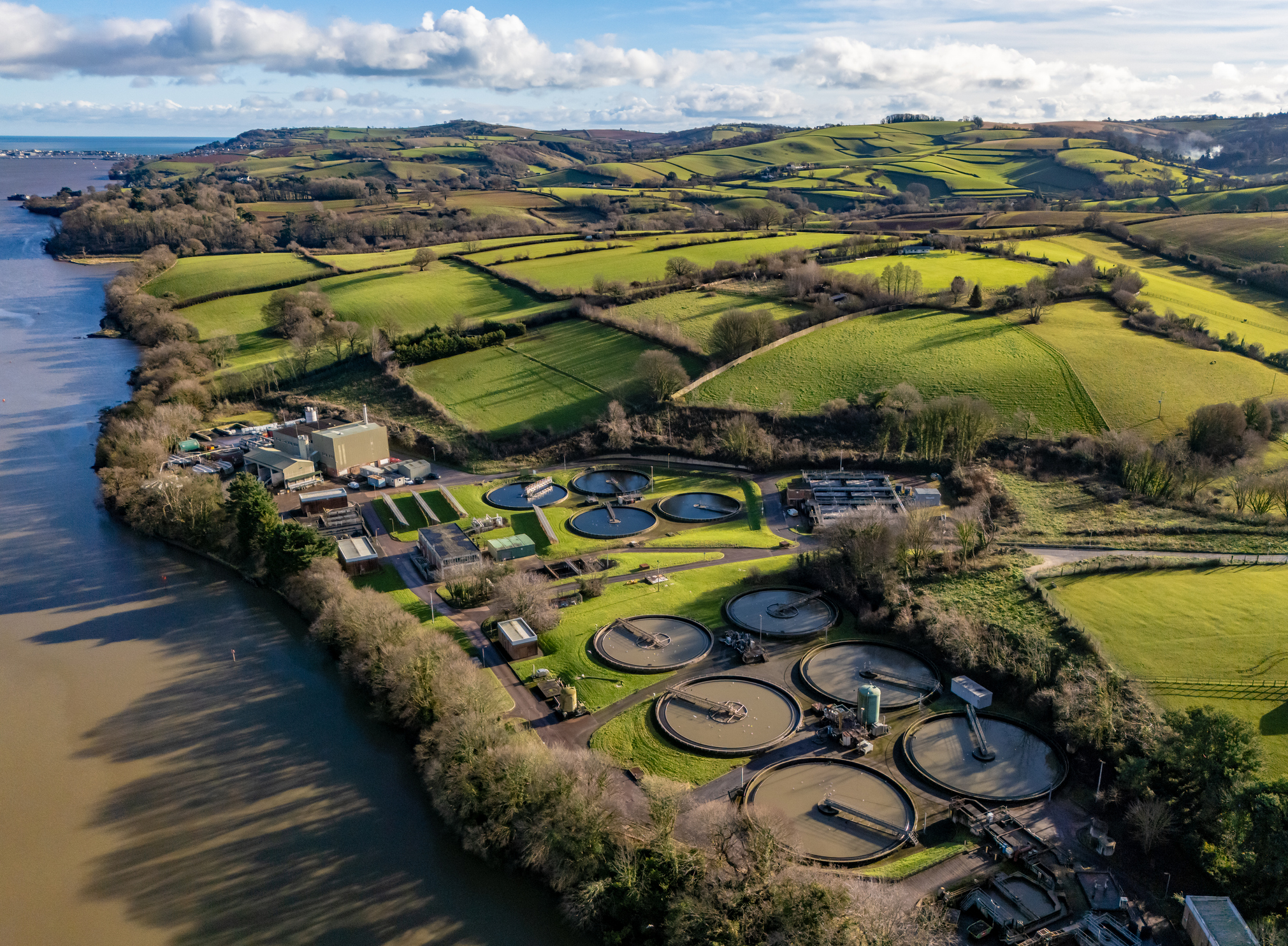 Aerial view of a water treatment plant with multiple circular clarifiers next to a river, surrounded by green fields and rolling hills under a partly cloudy sky.