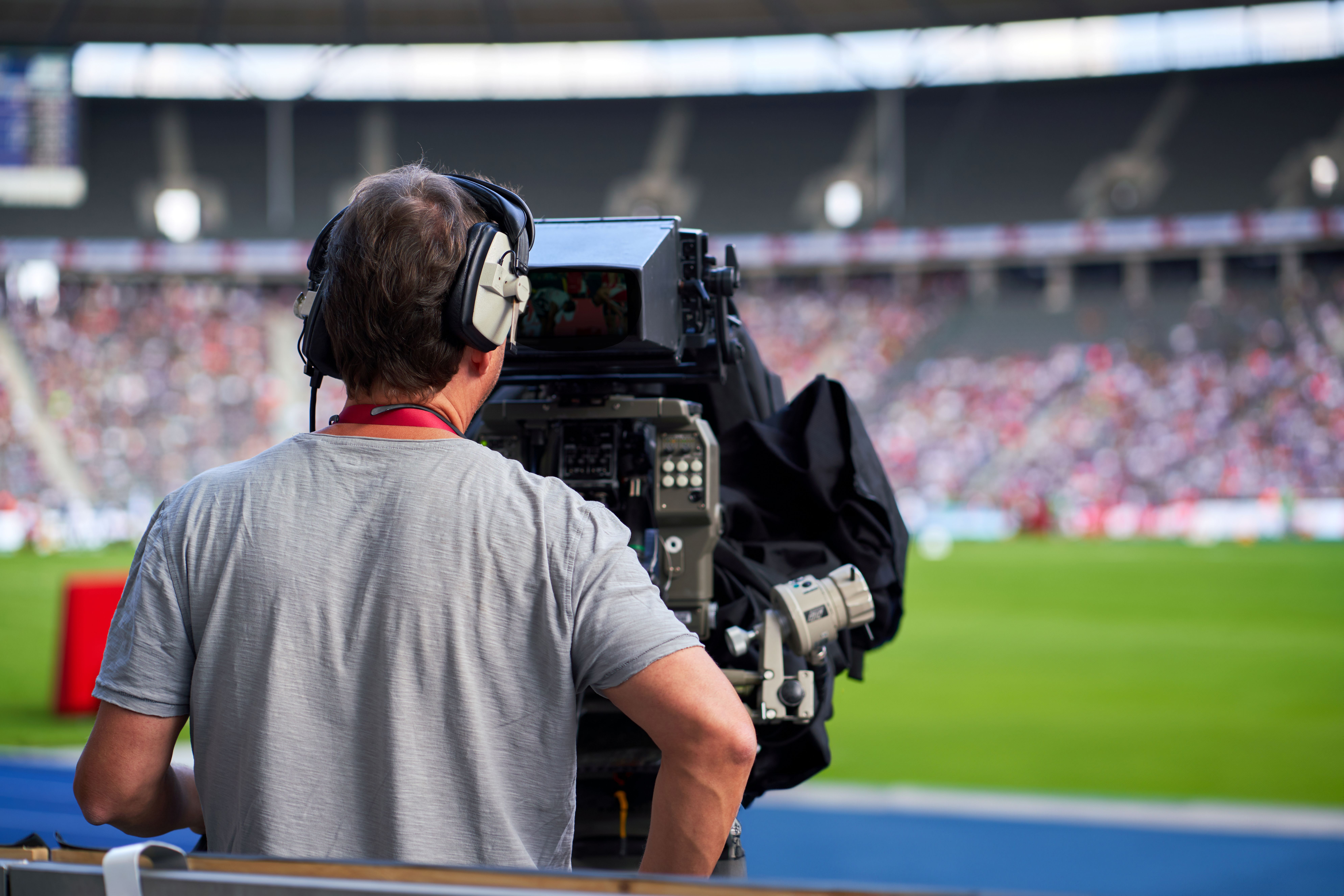 A cameraman wearing headphones filming a sports event in a stadium with a large crowd in the background.