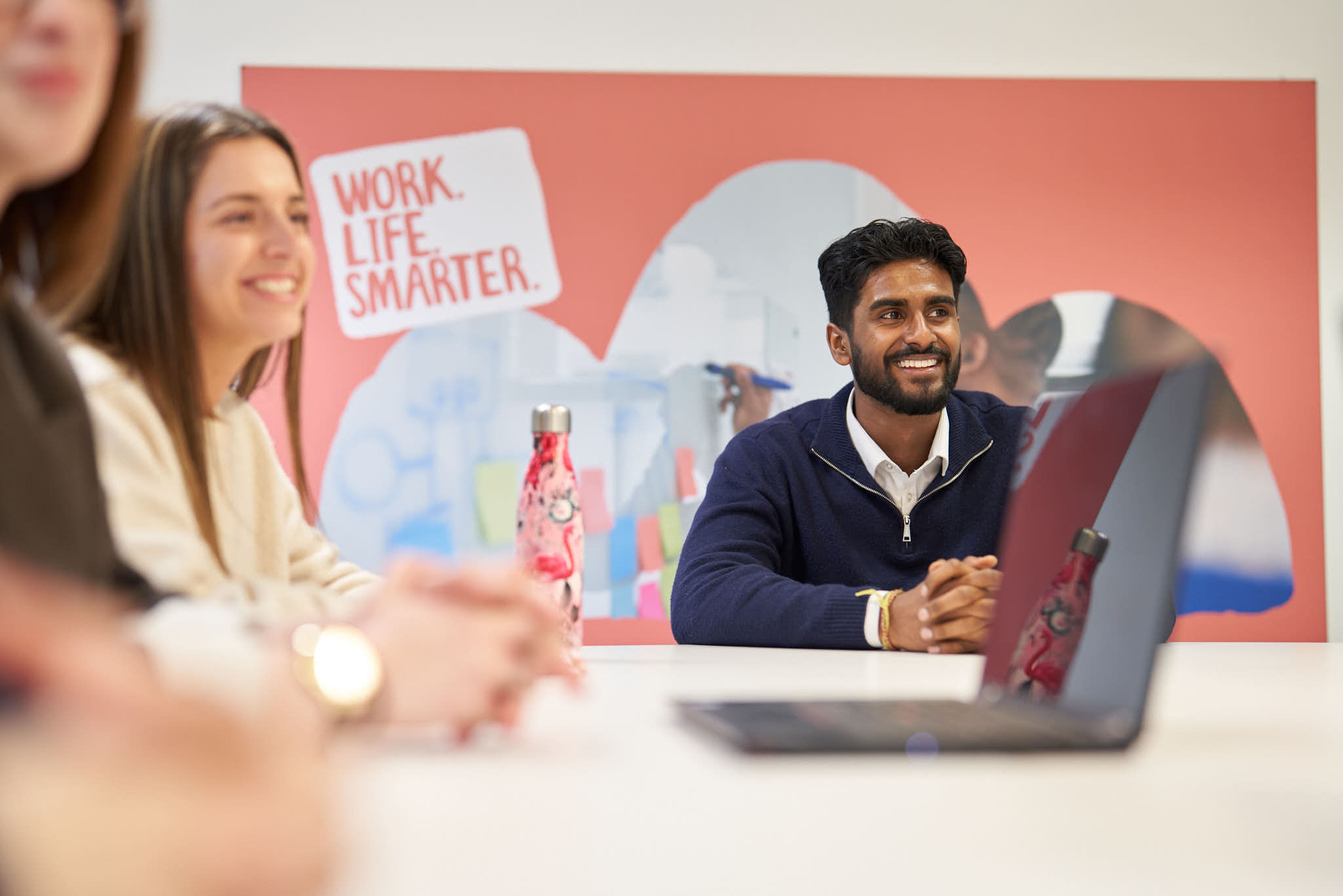 Smiling man with a beard sitting at a table during a meeting, with a blurred woman and hands in the foreground and a sign on the wall reading 'WORK. LIFE. SMARTER.'