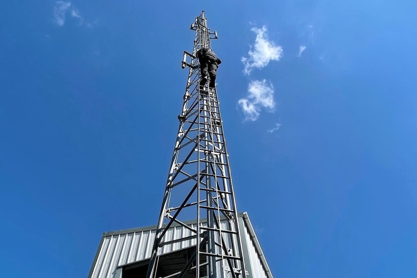A person in black climbing a tall metal communication tower against a clear blue sky with a few clouds, next to a corrugated metal building.