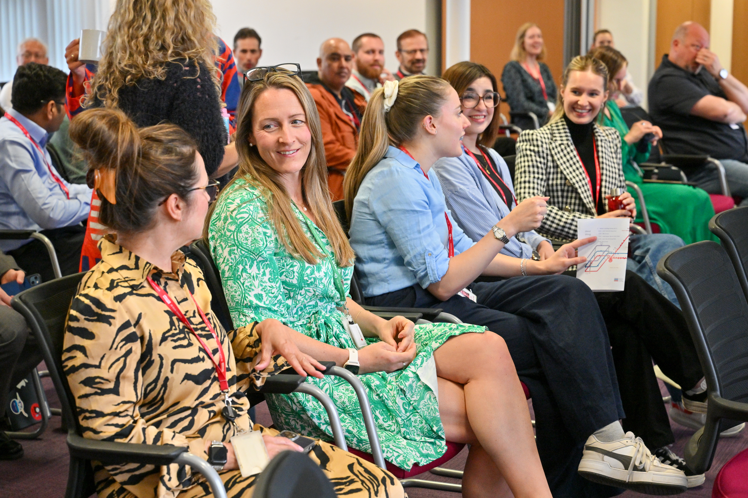 A group of people seated in a conference room, with several women in the foreground engaging in conversation and smiling, wearing name badges and casual to business-casual attire.