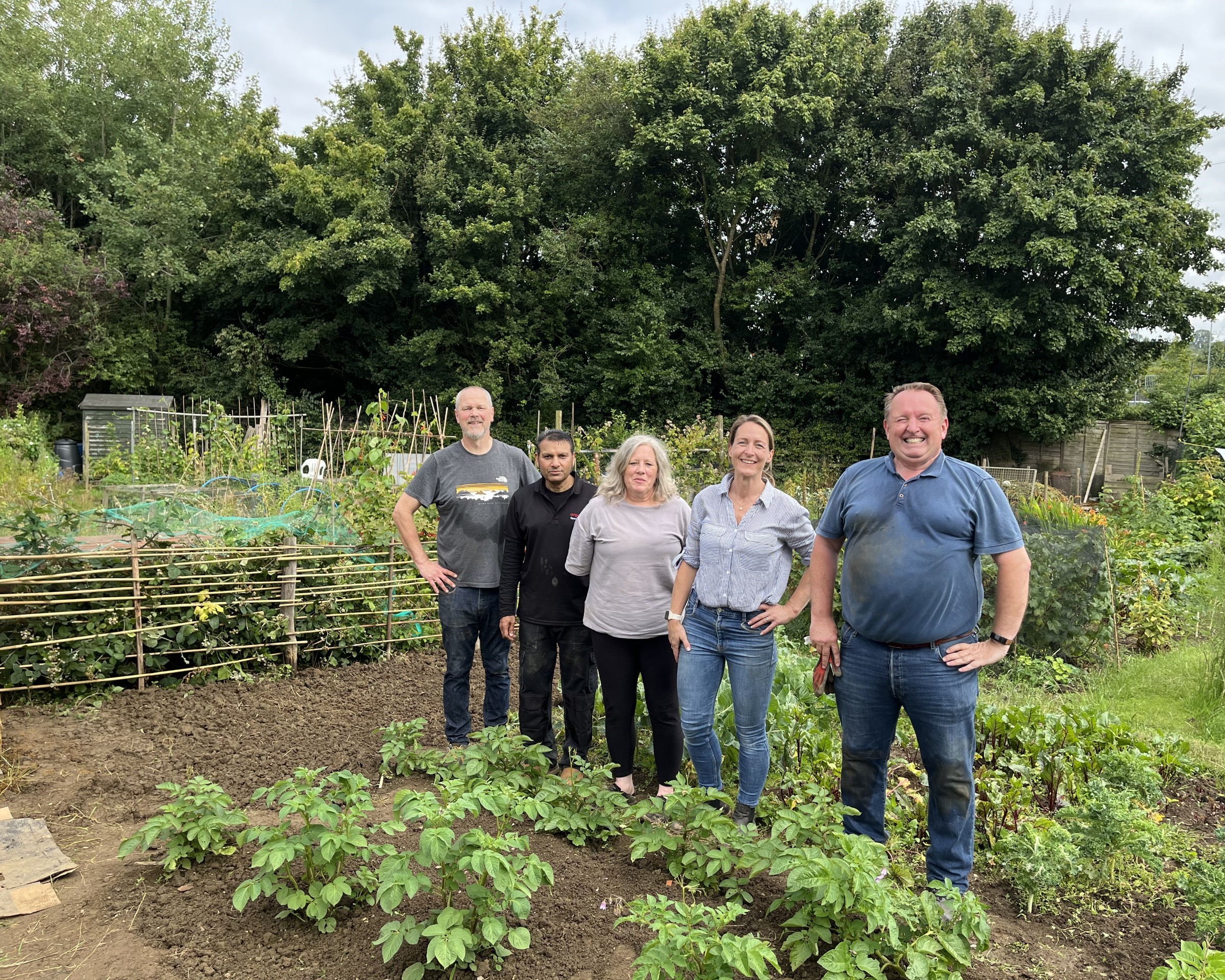 Five people standing side by side smiling in a community garden with green plants and trees in the background.
