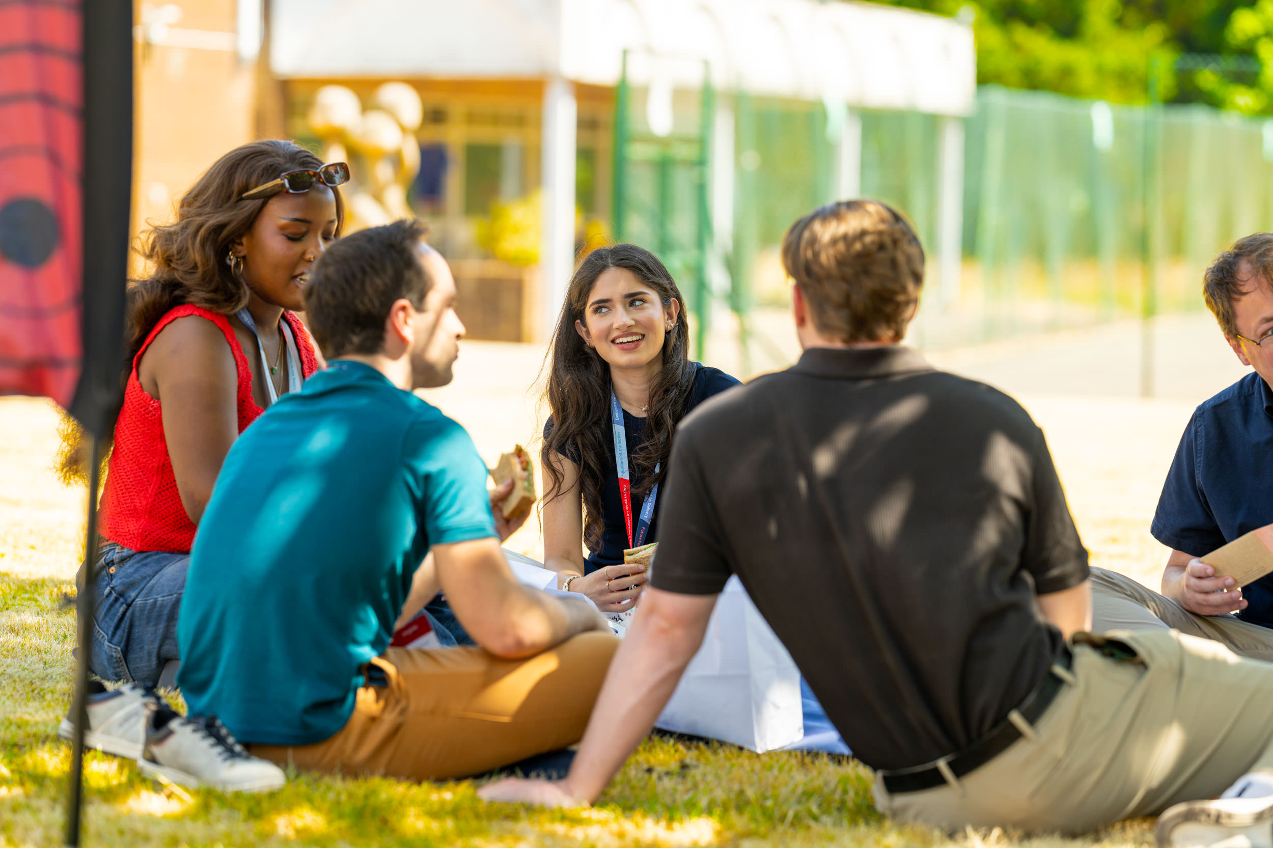 Five young adults sitting on grass outdoors, engaged in conversation and eating sandwiches, with a sunny background featuring a building and greenery.