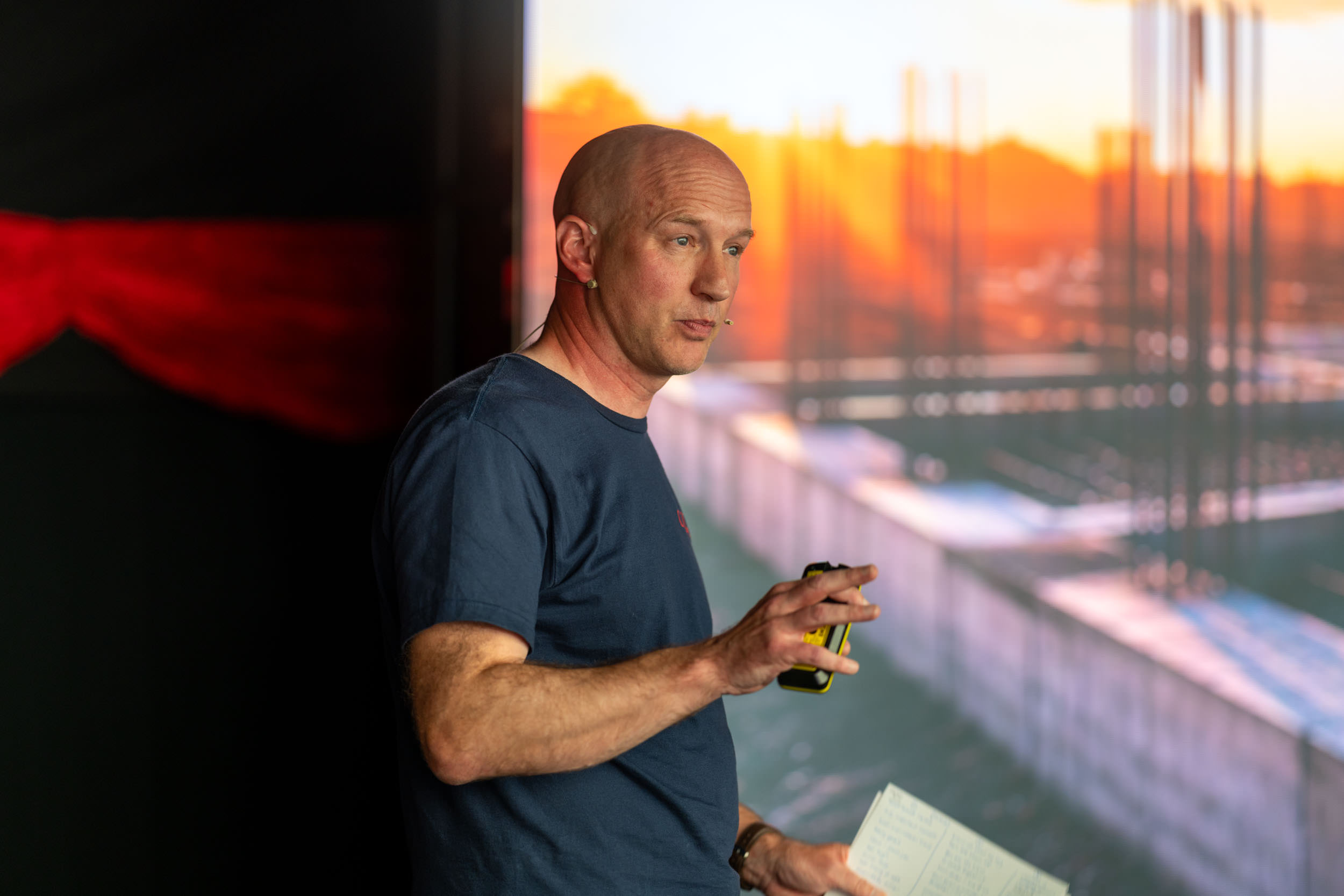 Dominic Biles wearing a navy blue t-shirt speaking with a headset microphone, holding a yellow and black device and notes, with a blurred sunset and outdoor scene in the background.