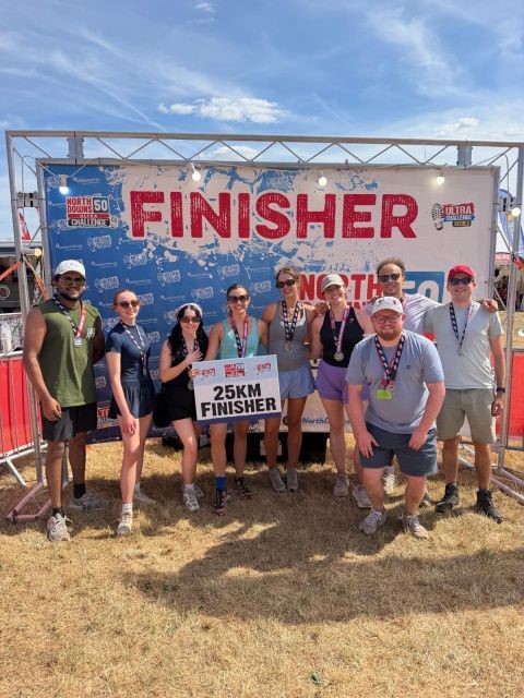 A group of nine people smiling and posing outdoors on dry grass in front of a large banner that reads 'FINISHER' and 'North Downs 50 Challenge.' One person in the center holds a sign saying '25KM FINISHER.' They are wearing casual athletic clothes and medals around their necks under a sunny sky.