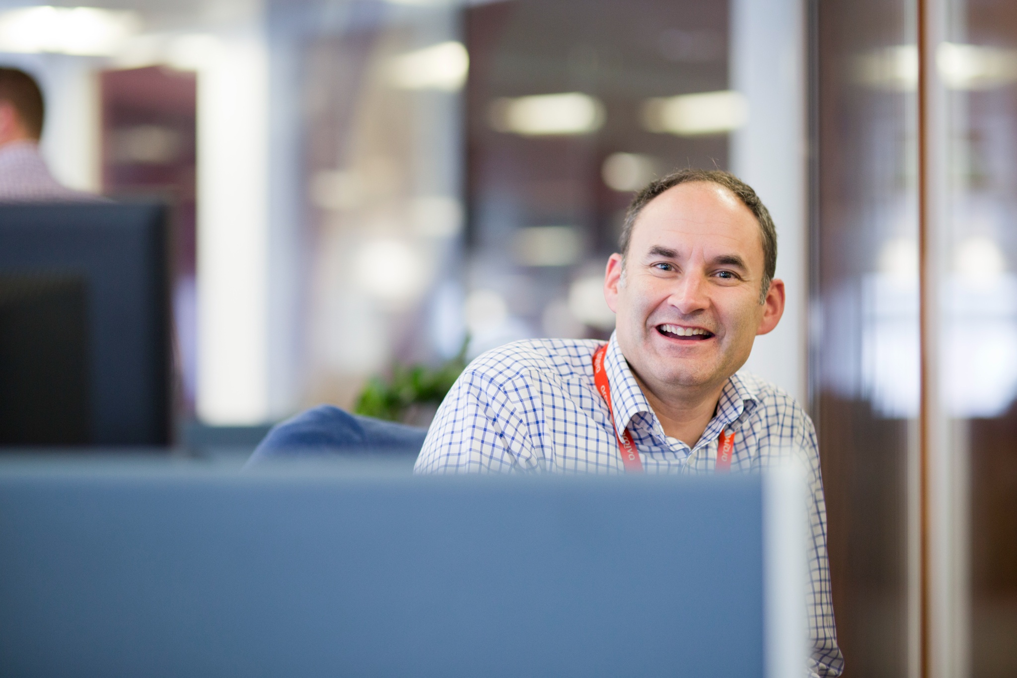 Smiling man wearing a checkered shirt and a red lanyard, sitting in a modern office cubicle.