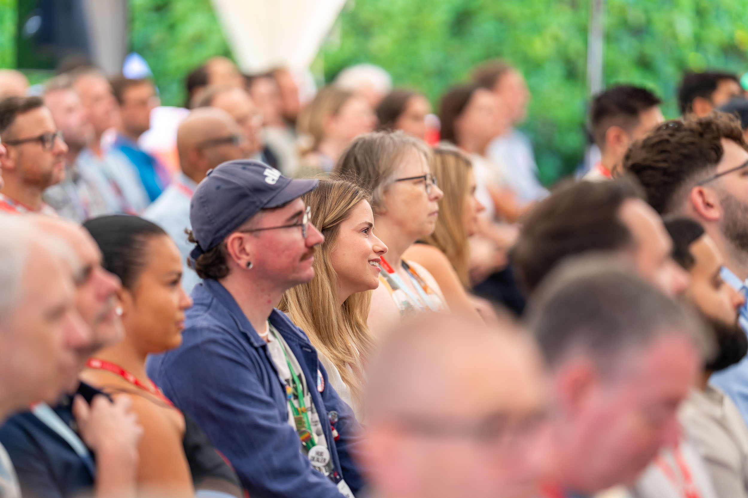 Audience of diverse adults seated in rows attentively listening at an outdoor event with green foliage in the background.