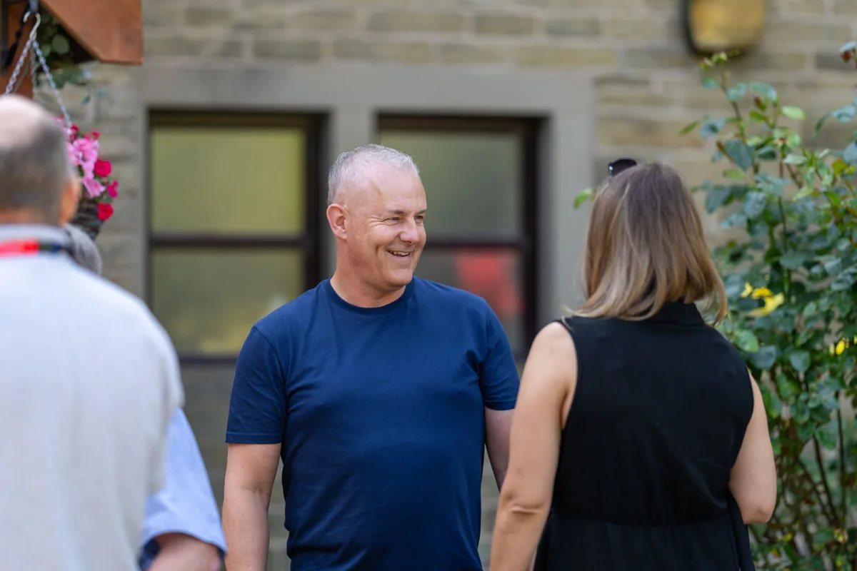 A man in a navy blue t-shirt smiling and talking to a woman in a black sleeveless top in an outdoor setting with a stone wall and window in the background.