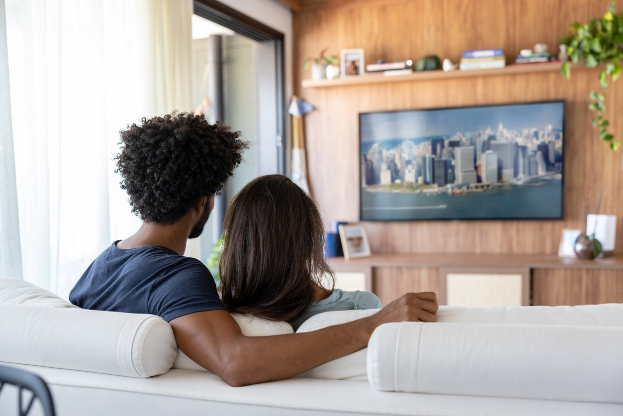 Couple sitting on a white couch in a living room, watching a city skyline image on a wall-mounted TV.