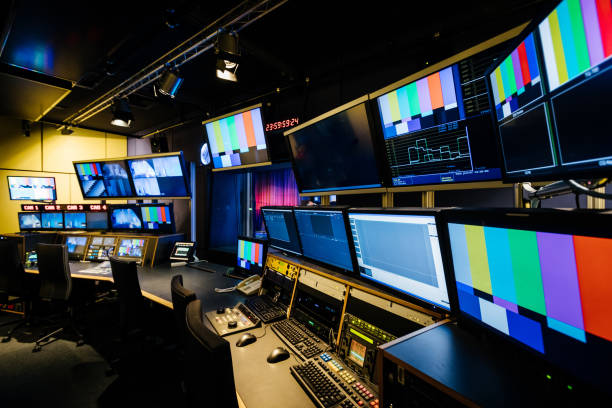 Broadcast control room with several monitors displaying color bars and waveform patterns, control panels with buttons and knobs, and empty chairs.