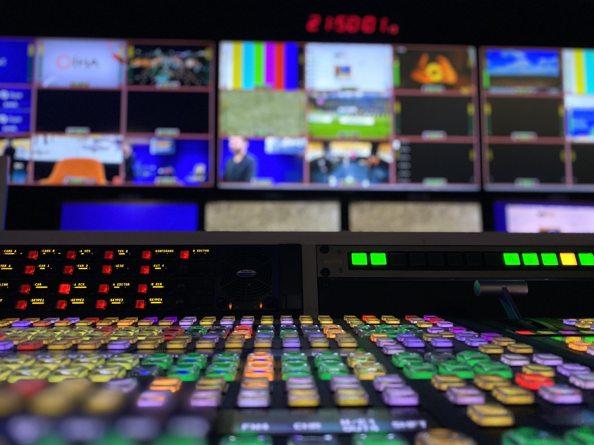 Close-up of a broadcast control panel with numerous colorful illuminated buttons and screens in the background showing multiple video feeds in a control room.