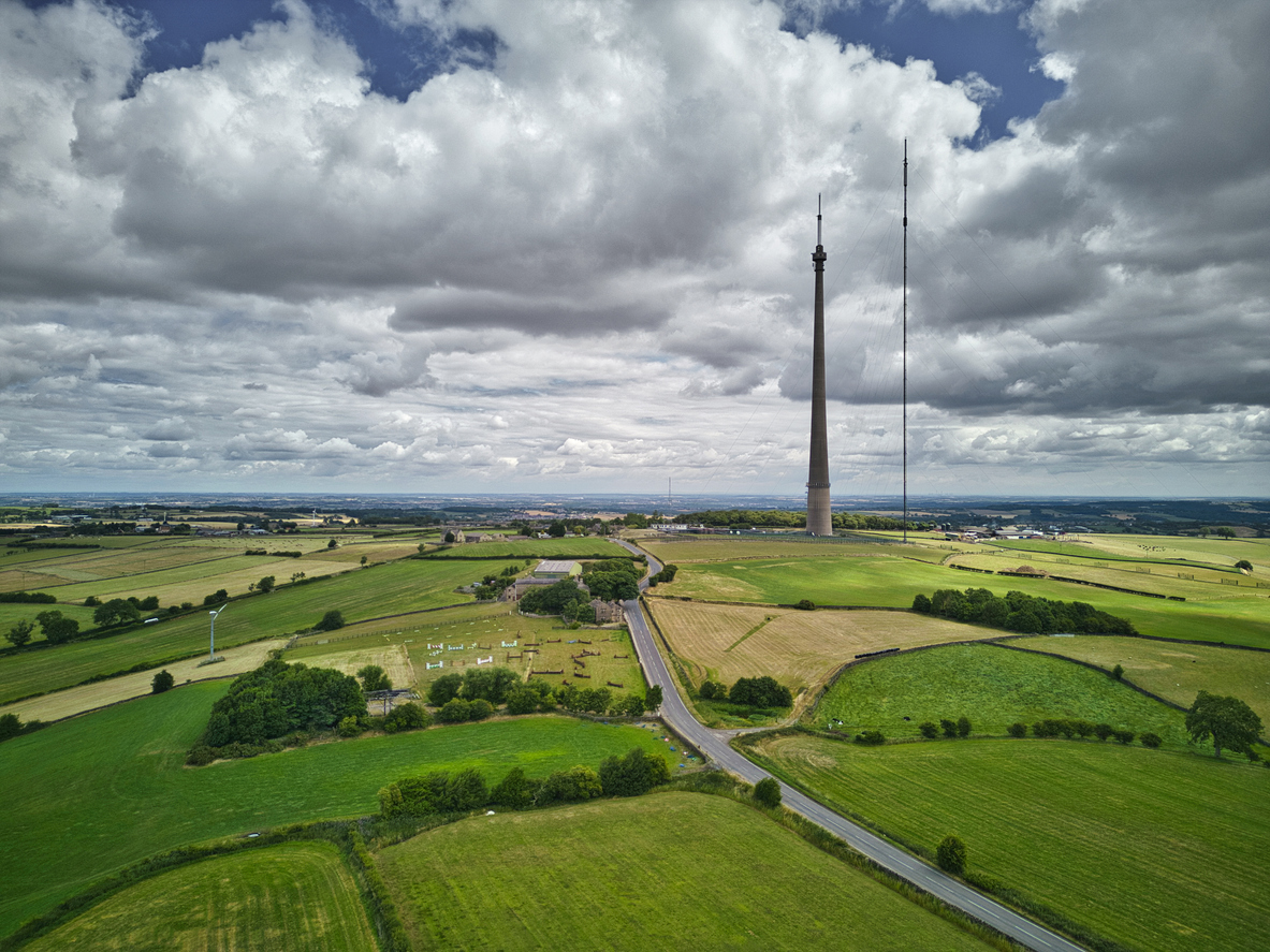 Aerial view of green fields and farmland with a narrow road running through them and two tall broadcast towers standing prominently under a cloudy sky.