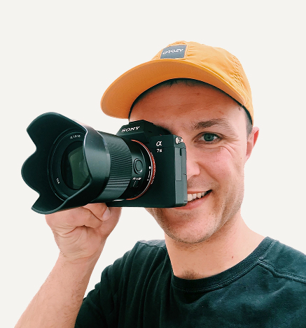 Smiling man wearing a yellow cap and black shirt holding a Sony Alpha 7 II camera up to his eye.