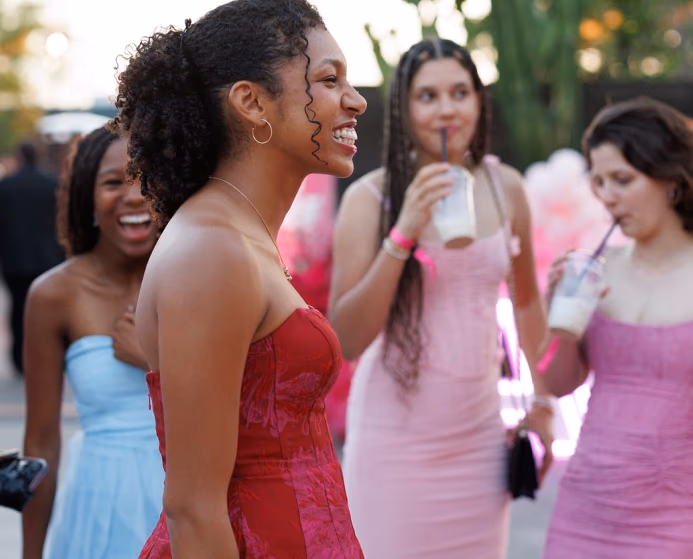 Four young women in colorful dresses smiling and drinking beverages outdoors at a social event.