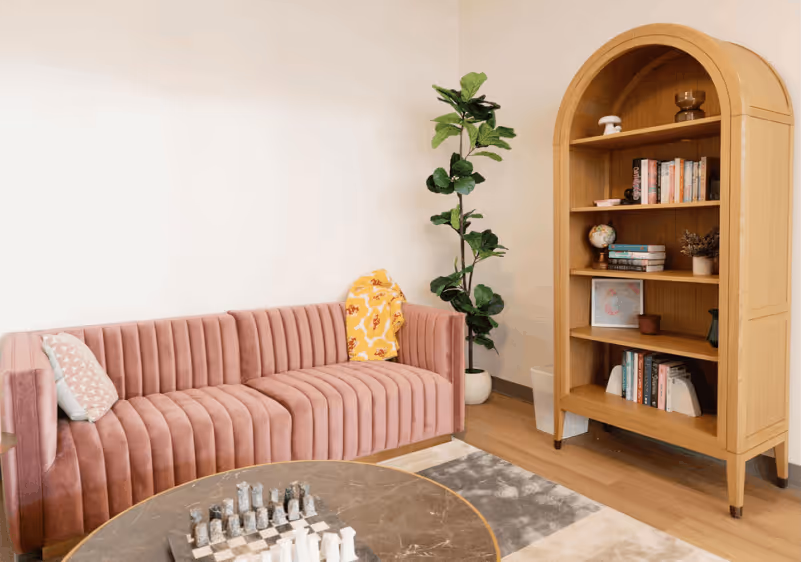 Modern living room with pink velvet sofa, tall green plant, wooden bookshelf, and chessboard on table.