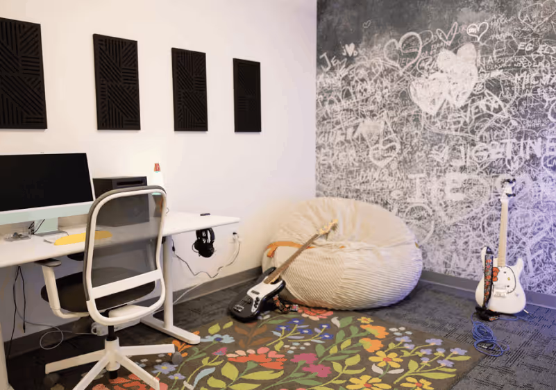 Modern room with computer desk, mesh chair, bean bag, two electric guitars, and floral rug on carpeted floor.