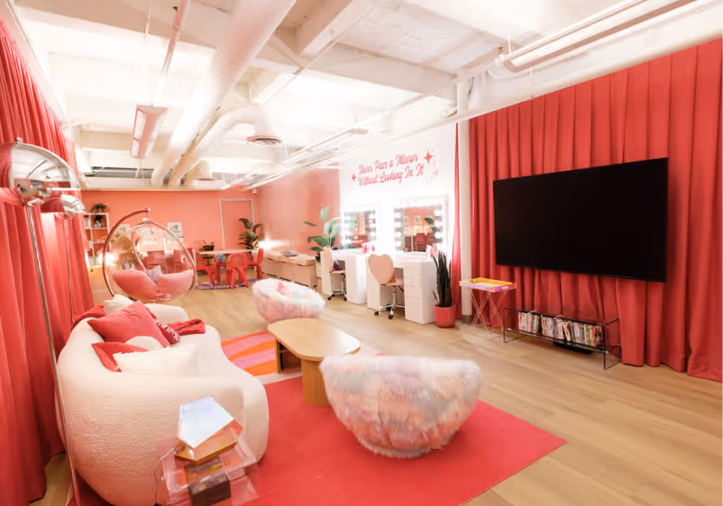 Bright pink-themed lounge with comfy chairs, vanity mirrors, a large screen TV, and books on a shelf.