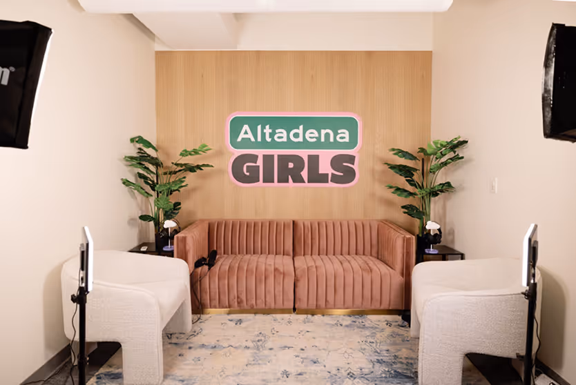 Room with pink couch, two white chairs, plants, and 'Altadena GIRLS' sign on wood wall.