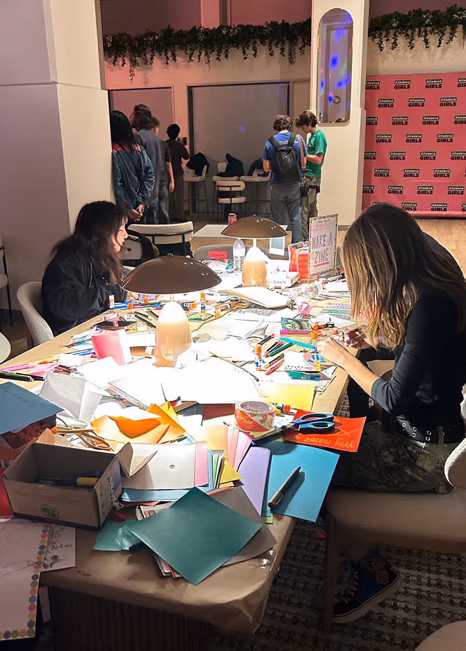Two women crafting with paper, glue, and markers at a cluttered table under lamps at an event space.